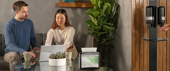 To som sitter på pauserom og en som kommer inn å bruker en antibakterie dispenser