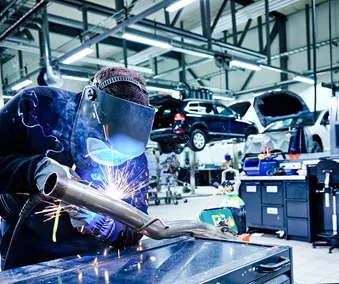 En person som sveiser med verneutstyr og ett førstehjelpsett i bakgrunnen