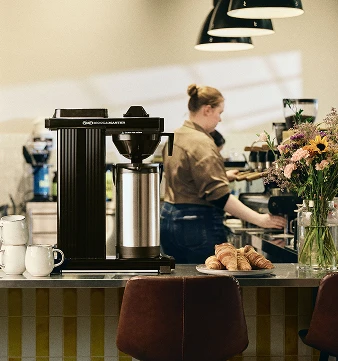 Bilde av en moccamaster med mennesker i bakgrunnen som holder på å lage kaffe