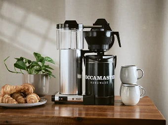 Bilde av en moccamaster på ett brunt bord med kaffekopper på en side croissant på den andre med en potteplante litt tilbake på den ene siden
