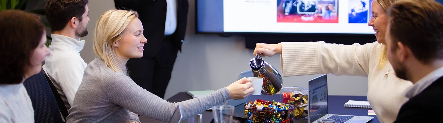 Bilde av noen mennesker på et møterom hvor en serverer kaffe til en annen