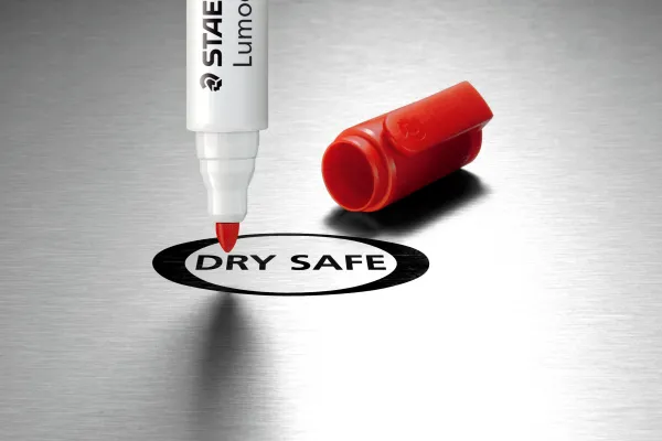 Rød Staedtler Lumocolor tusj som peker ned mote en metalisk overflate det står dry safe på, men rød kork liggende på overflaten