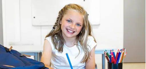 Ett barn som sitter ved skolebenken og ser mott kamera med en fargeblyant i hånden